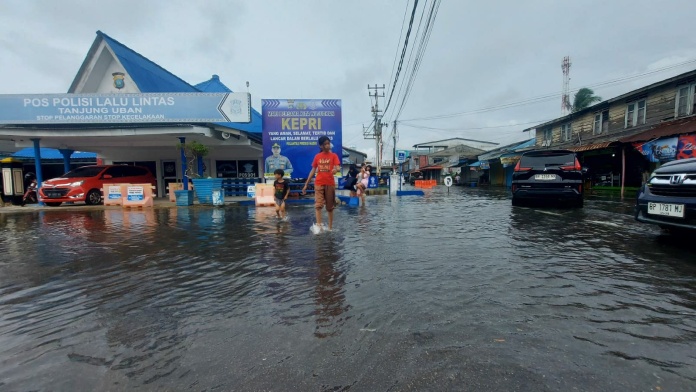Seorang warga berjalan melewati jalan yang tergenang banjir rob di Tanjung Uban, Kepulauan Riau. Di latar belakang, tampak Pos Polisi Lalu Lintas dan sejumlah kendaraan yang terparkir sebagian terendam air akibat banjir rob. Langit tampak mendung, menandakan cuaca ekstrem yang masih berpotensi terjadi.