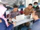 Sekelompok orang sedang melakukan proses administrasi di sebuah meja layanan publik. Seorang petugas berseragam polisi duduk di depan laptop, berhadapan dengan seorang wanita berhijab yang menyerahkan dokumen. Di sebelahnya, seorang petugas berseragam cokelat turut membantu di depan laptop lain, sementara seorang pria berpakaian biru duduk di sisi meja yang sama. Di belakang mereka, terlihat satu orang lagi mengenakan masker hijau dan pakaian batik, tampak sedang mengamati proses tersebut. Latar belakang menunjukkan papan dengan logo 